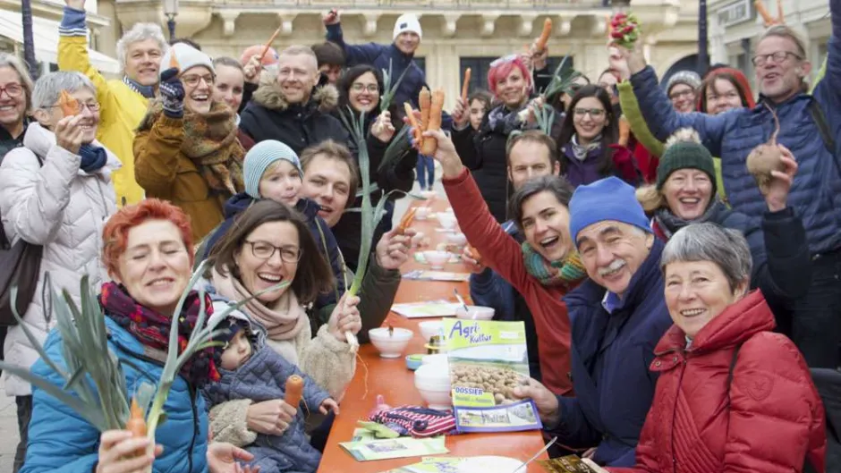 Zu sehen ist ein mittig aufgestellter langer Tisch, dan dem viele fröhliche Menschen jeden Alters sitzen und teils Gemüse in die Luft halten.